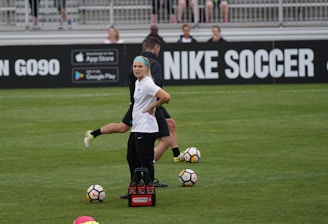 A person in sports attire stands on a soccer field, surrounded by several soccer balls and a carrier with bottles. The field is bordered by advertisements for Nike Soccer and app stores. Several people sit in bleachers in the background.