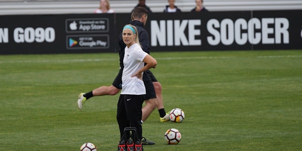 A person in sports attire stands on a soccer field, surrounded by several soccer balls and a carrier with bottles. The field is bordered by advertisements for Nike Soccer and app stores. Several people sit in bleachers in the background.