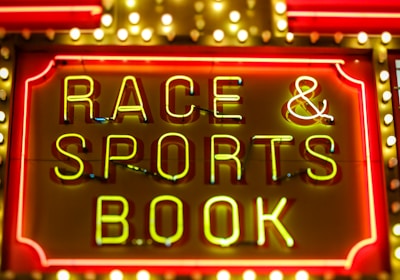 Bright neon lights on a marquee sign display the words 'RACE & SPORTS BOOK'. The sign is surrounded by rows of yellow and white lights against a red background, evoking a vibrant, energetic atmosphere typically associated with casinos or entertainment venues.