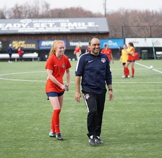 A group of people are on a soccer field, with some wearing sports attire. In the foreground, a person in red sports clothing is walking alongside someone in a dark tracksuit. Other people are visible in the background, some wearing yellow jerseys. A building with advertising banners, including a prominent sign reading 'Ready, Set, Smile,' is in the background.