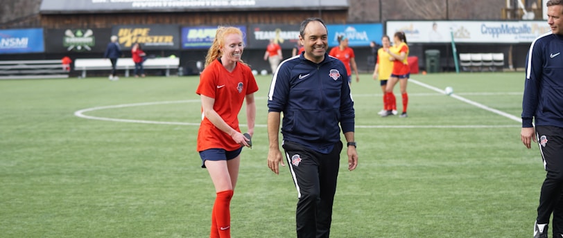 A group of people are on a soccer field, with some wearing sports attire. In the foreground, a person in red sports clothing is walking alongside someone in a dark tracksuit. Other people are visible in the background, some wearing yellow jerseys. A building with advertising banners, including a prominent sign reading 'Ready, Set, Smile,' is in the background.