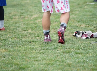 A person wearing sports shoes and patterned shorts stands on a grassy field, with various sports equipment like gloves and a helmet placed on the ground nearby.