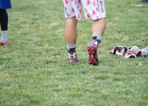 A person wearing sports shoes and patterned shorts stands on a grassy field, with various sports equipment like gloves and a helmet placed on the ground nearby.
