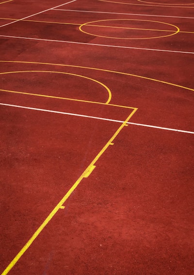 A sports court with a reddish-brown surface and bright yellow and white lines. The lines form geometric shapes, including rectangles and circles, suggesting markings for games like basketball or netball.