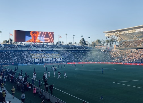 A large sports stadium filled with spectators. The field is occupied by soccer players, some walking and others scattered across the pitch. A massive screen displays a person, and flags fly on top of the stadium roof. The audience is dense, and the atmosphere seems lively with a visible banner showing 'BLUE WHITE & GOLD'.