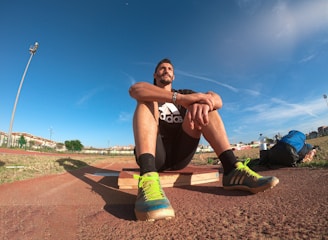 A person wearing a black athletic outfit is seated on a wooden platform in an outdoor sports field. The individual is relaxed, with arms resting on knees. The field is expansive with a clear blue sky and scattered clouds above. A brightly colored running shoe and athletic gear are visible, along with a water bottle and a blue backpack placed beside the person. Lampposts and residential buildings can be seen in the background.