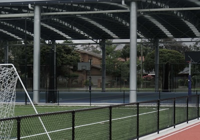 An outdoor sports facility featuring a running track and a soccer goal. The track is red, surrounded by a black metal fence, and the field is covered with artificial green turf. In the background, a basketball court is visible under a covered roof with metal supports. The sky is overcast, adding a moody atmosphere.