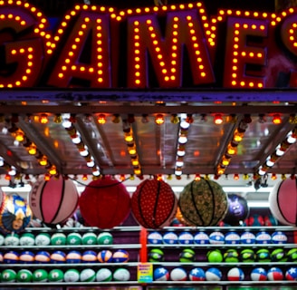 A brightly lit carnival game stall with a large 'GAMES' sign at the top surrounded by red and yellow lights. Various sports balls, including basketballs and soccer balls, are hanging from the ceiling and neatly arranged on shelves below.