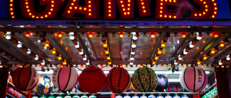 A brightly lit carnival game stall with a large 'GAMES' sign at the top surrounded by red and yellow lights. Various sports balls, including basketballs and soccer balls, are hanging from the ceiling and neatly arranged on shelves below.